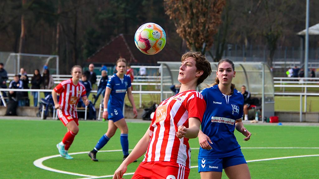 A female soccer player with short hair in a red jersey heads a ball on a green field. Two opponents are in the background.