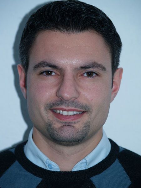 Ein Mann mit dunklem Haar und einem leichten Bart lächelt in die Kamera, trägt einen Pullover mit V-Ausschnitt und ein Hemd.