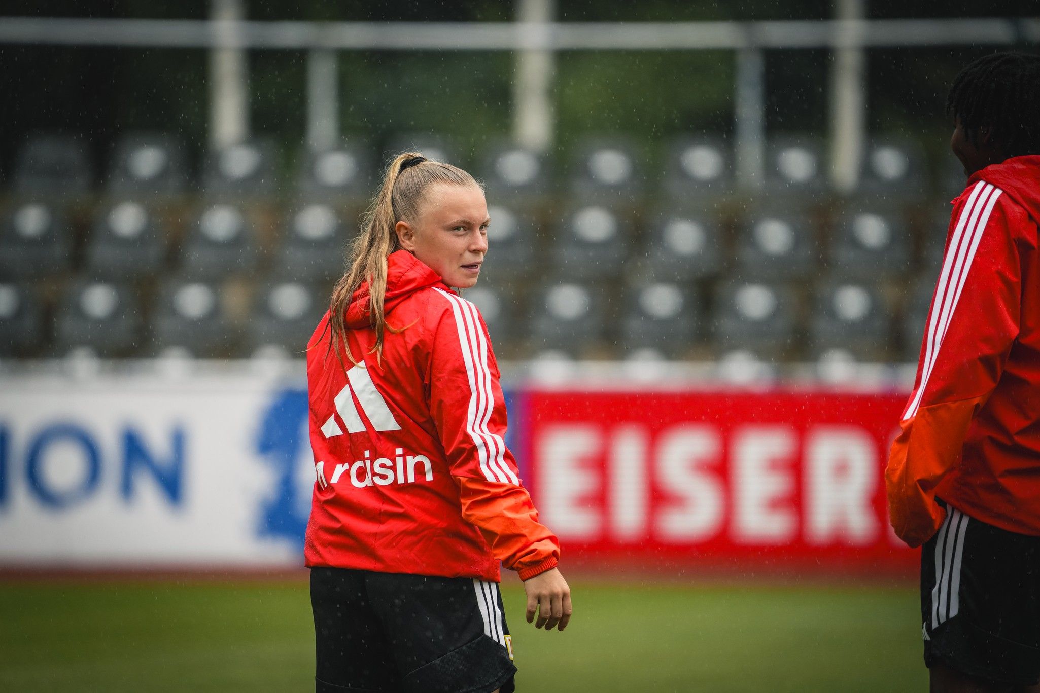 Eine Sportlerin in roter Trainingsjacke mit Adidas-Logo blickt während eines Trainings in die Kamera, im Hintergrund sind leere Sitze.