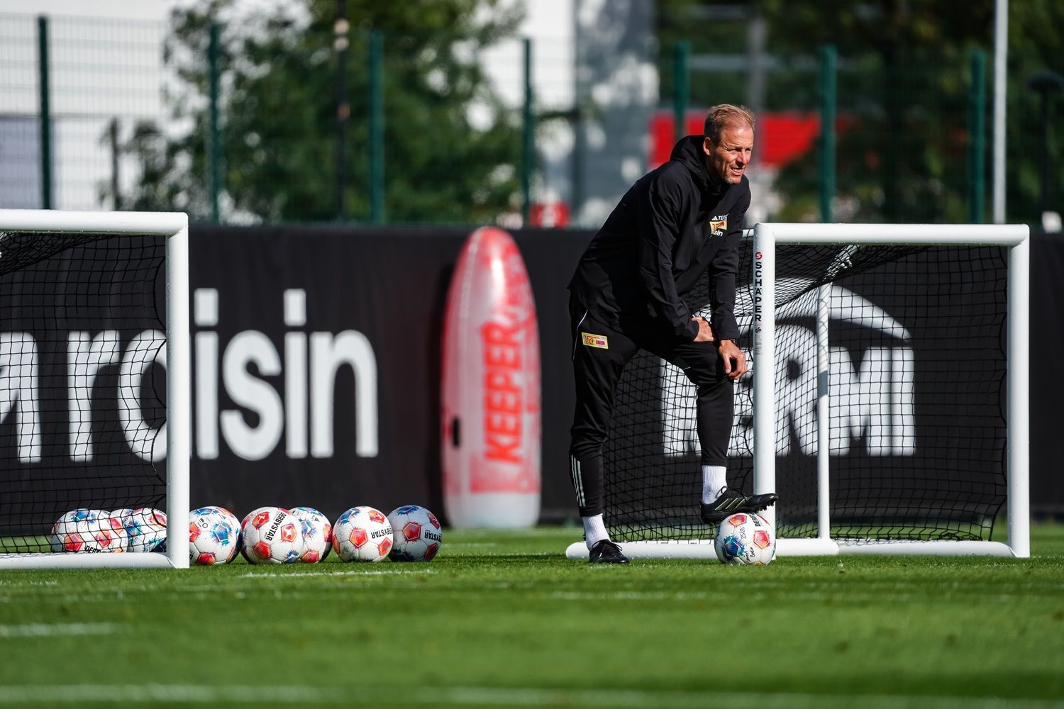 Ein Trainer steht neben einem Fußballtor und bereitet sich auf ein Training vor, während Bälle im Hintergrund liegen.