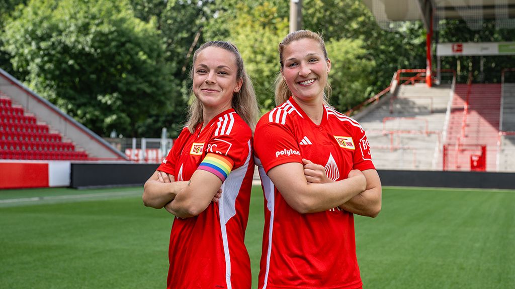 Two women stand on a soccer field, wearing red jerseys and displaying confidence with their arms crossed.