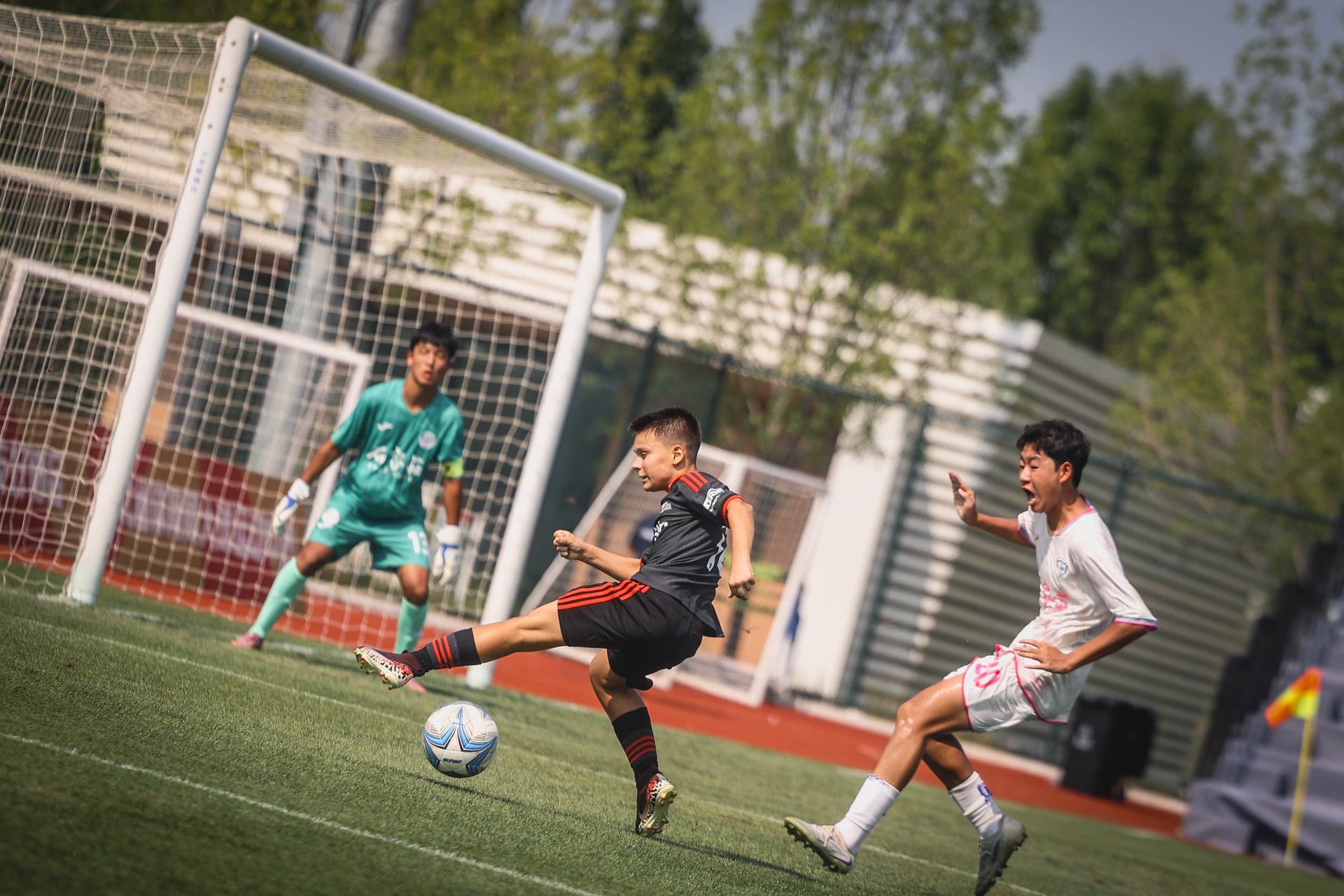 Ein junger Fußballspieler schießt den Ball, während ein gegnerischer Spieler in der Nähe ist und ein Torwart bereit steht.