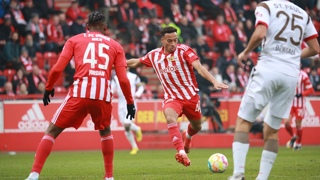 Two football players in red jerseys are fighting for the ball while an opponent in white is chasing them. Stadium atmosphere in the background.