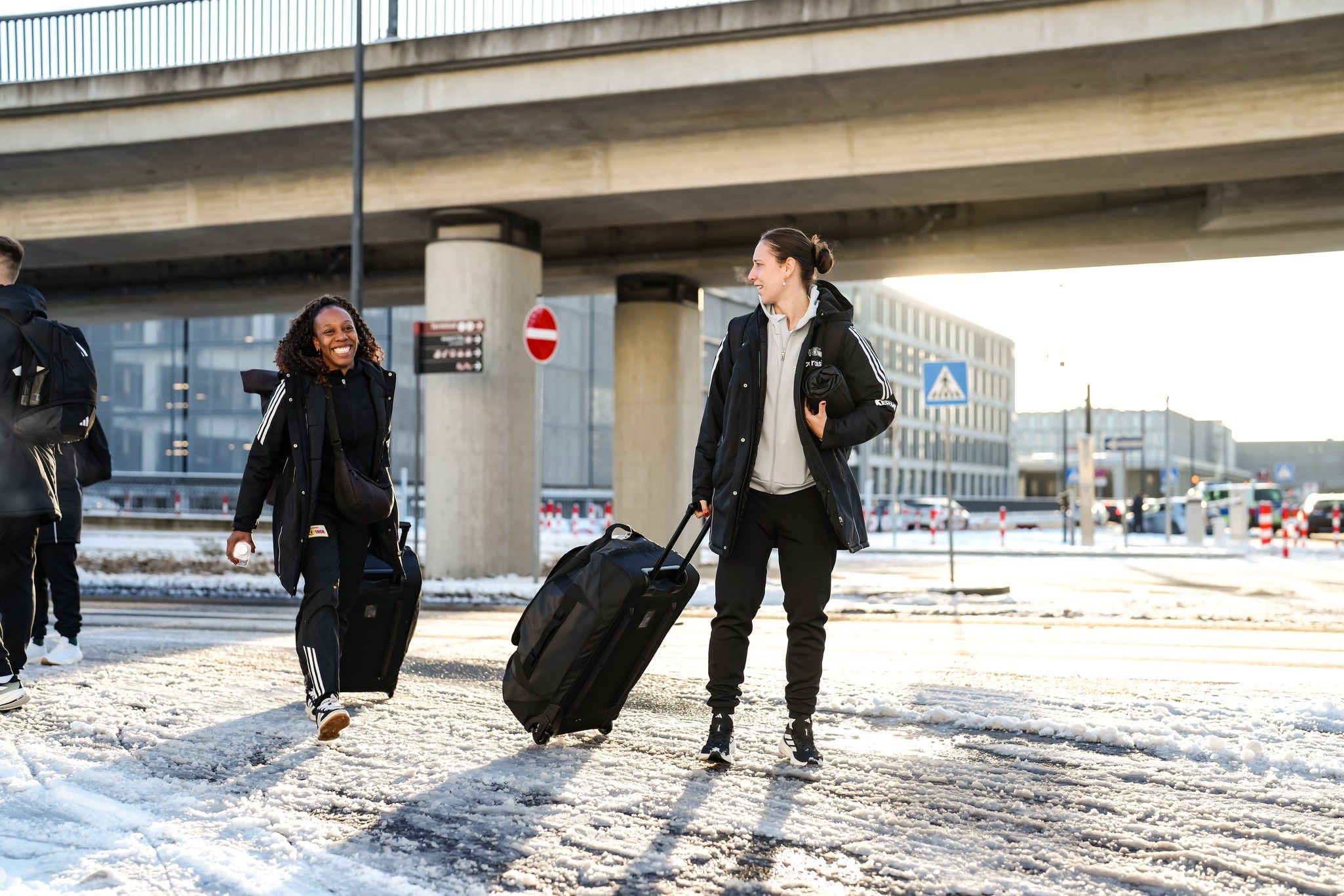 Zwei Personen mit Koffern gehen auf einer schneebedeckten Straße unter einer Brücke entlang.