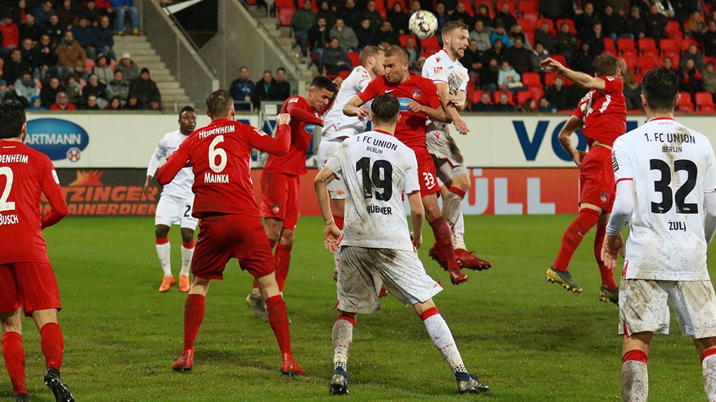 Es gibt ein Fußballspiel. Die Spieler tragen rot-weiße Trikots. Sie kämpfen um den Ball. Im Hintergrund stehen Zuschauer.