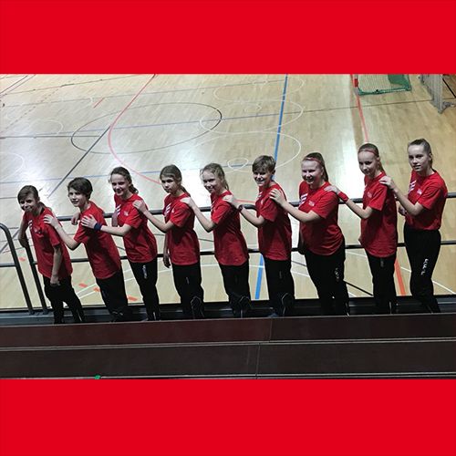 Eine Gruppe von zehn Jugendlichen in roten T-Shirts steht auf einer Tribüne und lächelt, während sie sich gegenseitig am Schulterblatt fassen.