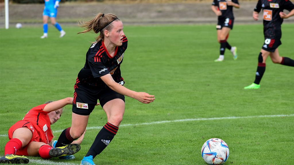Eine Spielerin in einem schwarzen Fußballtrikot sprintet mit dem Ball über das Spielfeld, während eine andere Spielerin am Boden liegt.