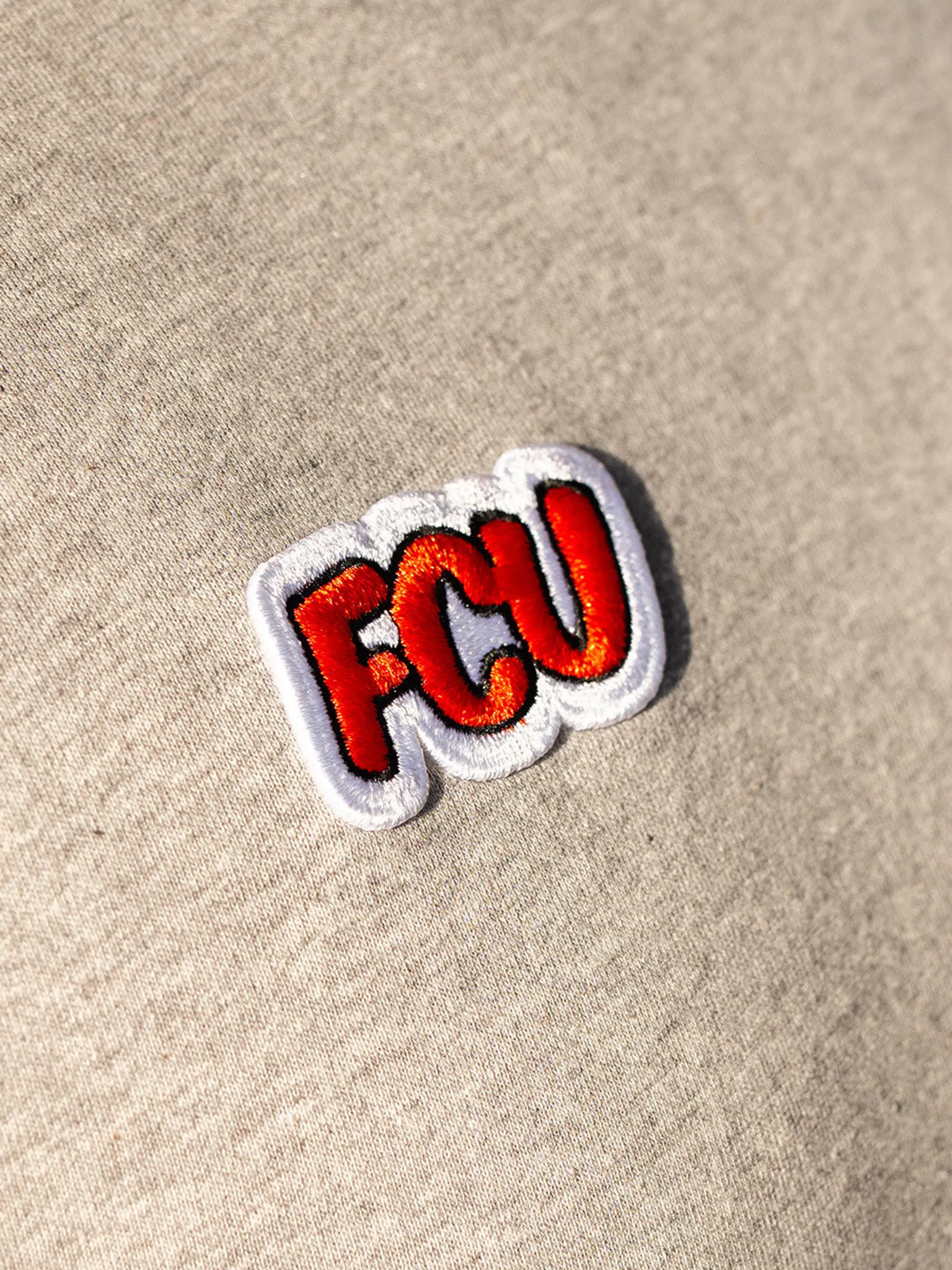 Ein aufgenähtes, farbiges Patch mit den Buchstaben "FCU" auf einem hellen, einfarbigen Stoff.