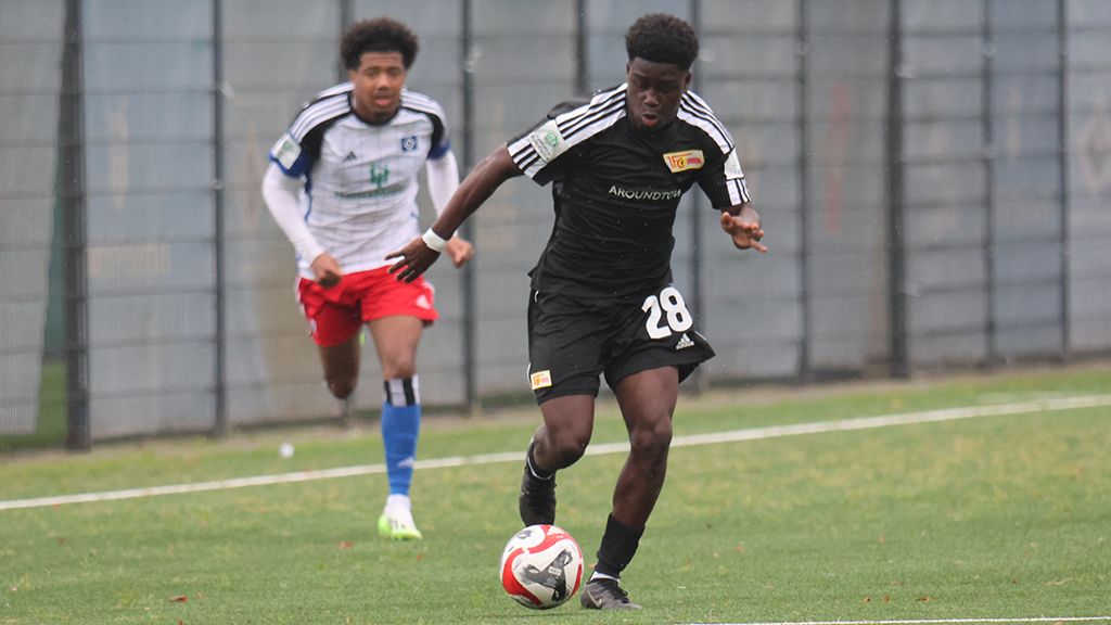 Ein Fußballer in schwarzem Trikot mit der Nummer 28 dribbelt den Ball, während ein Gegner in blau-weißer Kleidung folgt.
