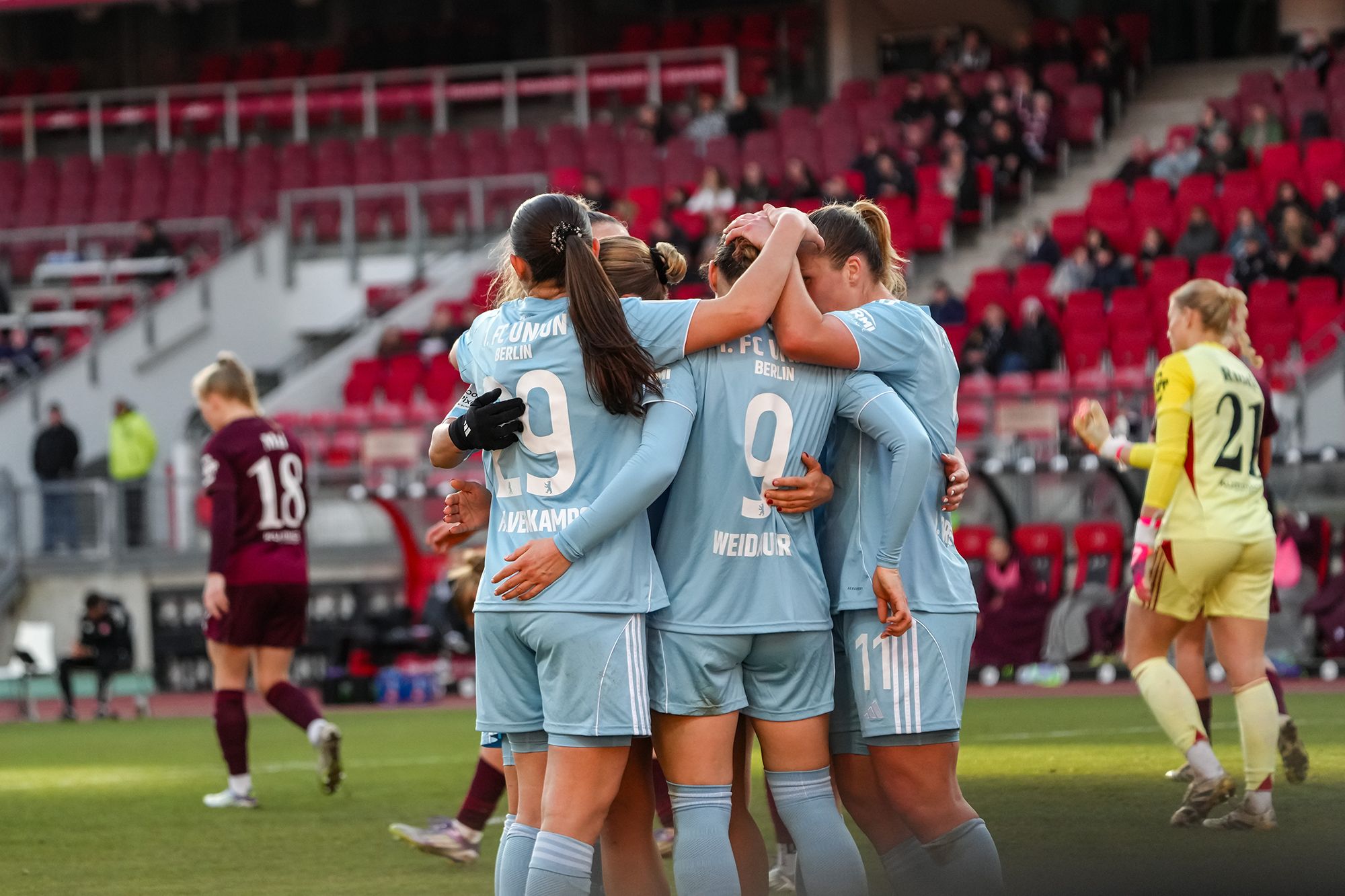 Fünf Fußballspielerinnen in blauen Trikots umarmen sich jubelnd auf dem Spielfeld während eines Spiels.