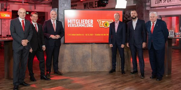 Six gentlemen in suits stand in front of a screen that says "Members Meeting." Background in a modern event space.