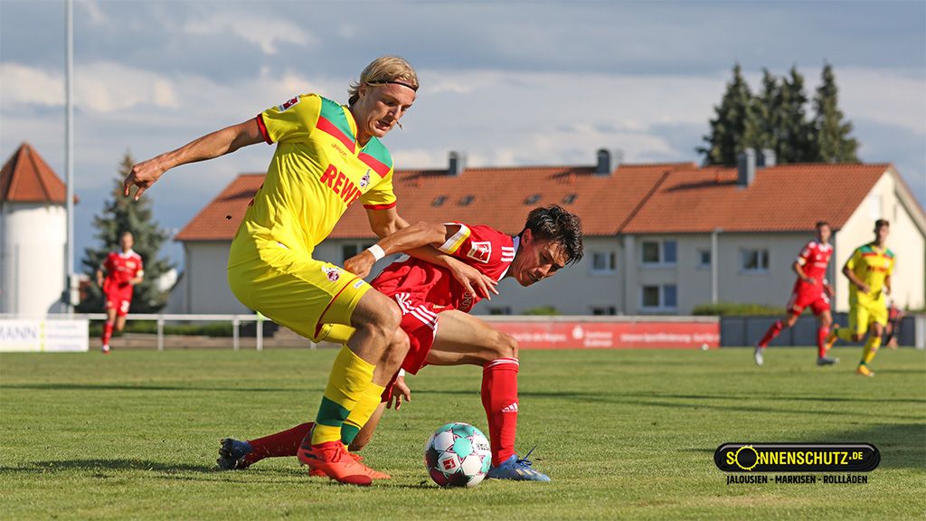 Zwei Fußballspieler in konkurrierender Position auf einem Rasenfeld, einer fällt, während der andere ihn drängt.