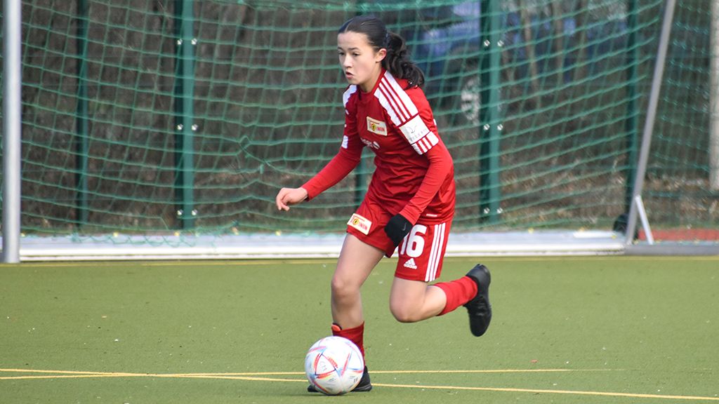 Mädchen in rotem Fußballtrikot spielt auf einem Platz und dribbelt den Ball.