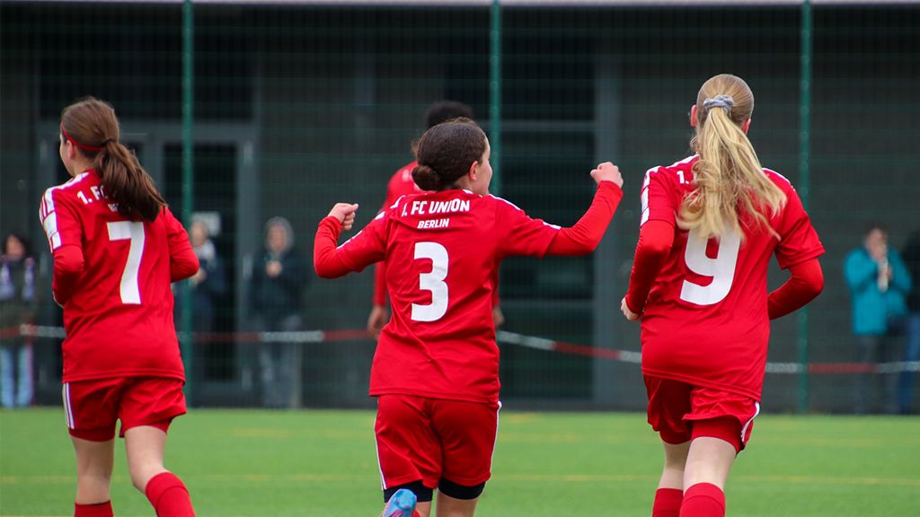 Mädchenfußballmannschaft in roten Trikots, die jubelnd auf dem Spielfeld läuft.