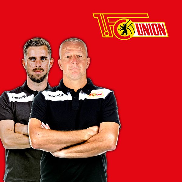 Zwei Männer in schwarz-roten Polo-Shirts stehen vor einem roten Hintergrund mit dem Logo des 1. FC Union Berlin.