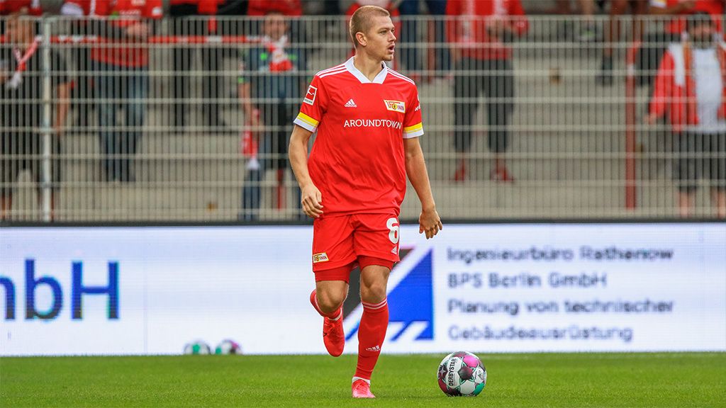 Ein Fußballspieler in rotem Trikot steht auf dem Spielfeld, umgeben von Zuschauern in einem Stadion.