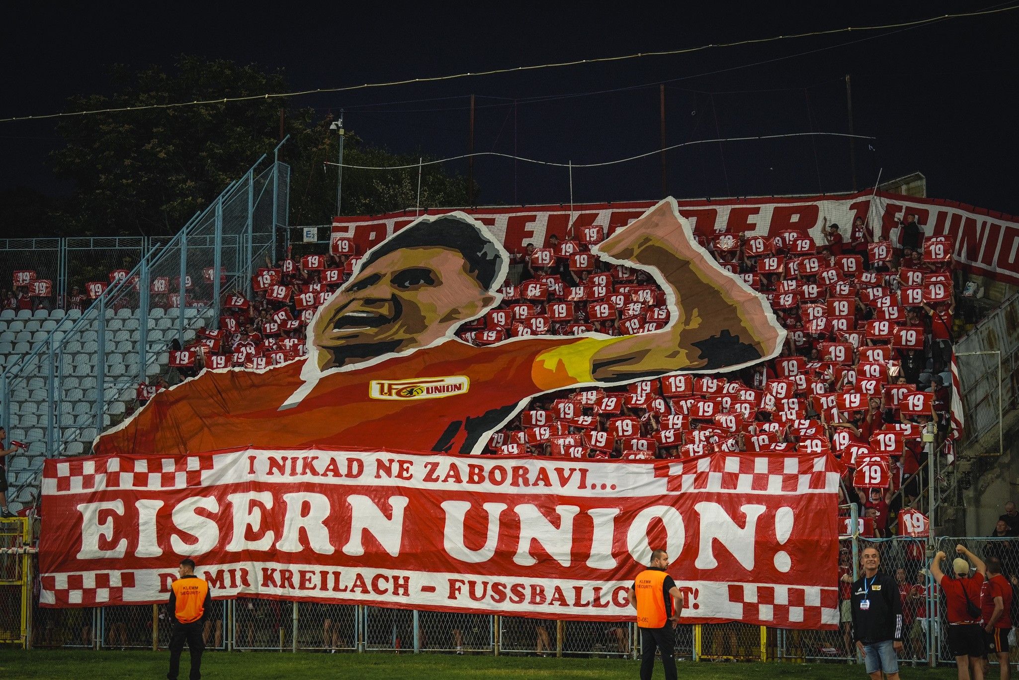 Große Fankurve mit Banner und vielen Fans in roten T-Shirts, die einen Fußballverein unterstützen.