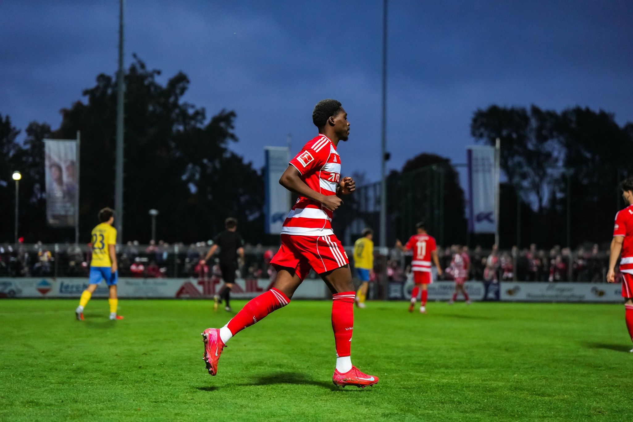 A football player in red and white kit jogs on the field, surrounded by spectators and other players at dusk.