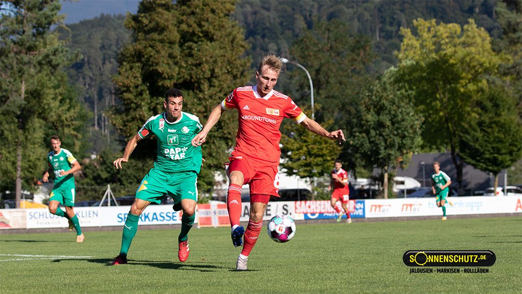 Ein Fußballspieler in roter Kleidung dribbelt den Ball, während ein Gegner in grüner Kleidung ihn verfolgt. Hintergrund mit Bäumen.