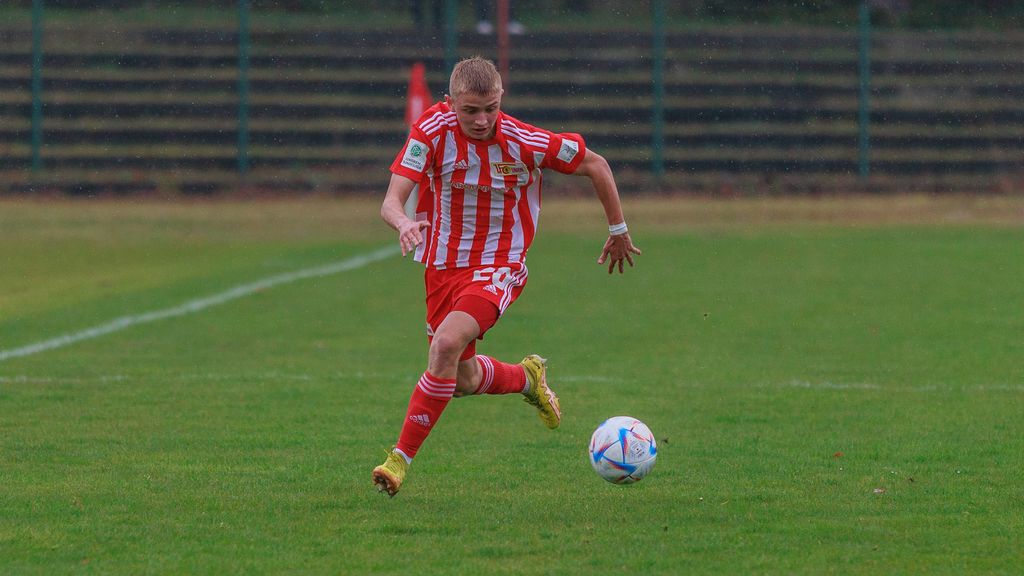 Ein junger Fußballspieler in rot-weißem Trikot läuft konzentriert über ein grünes Spielfeld.