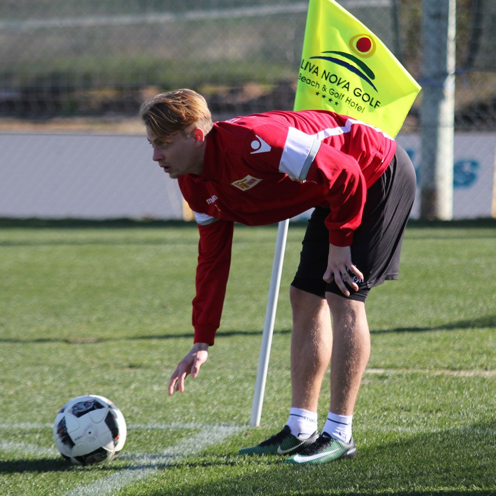 Ein Spieler in rotem Trikot beugt sich über einen Fußball, während er sich in der Nähe eines Eckfahne befindet.