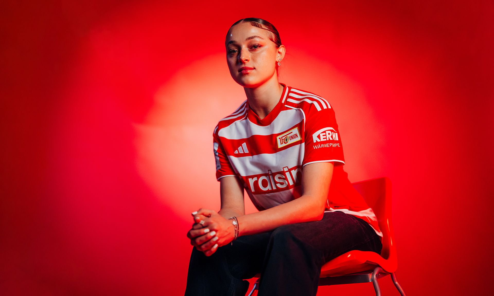 Eine Person sitzt auf einem roten Stuhl, trägt ein gestreiftes Fußballtrikot und hat einen neutralen Gesichtsausdruck vor einem roten Hintergrund.