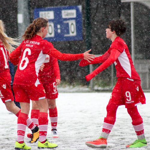Mädchenfußballteam in roten Trikots feiert bei Schneefall einen Torerfolg auf dem Spielfeld.