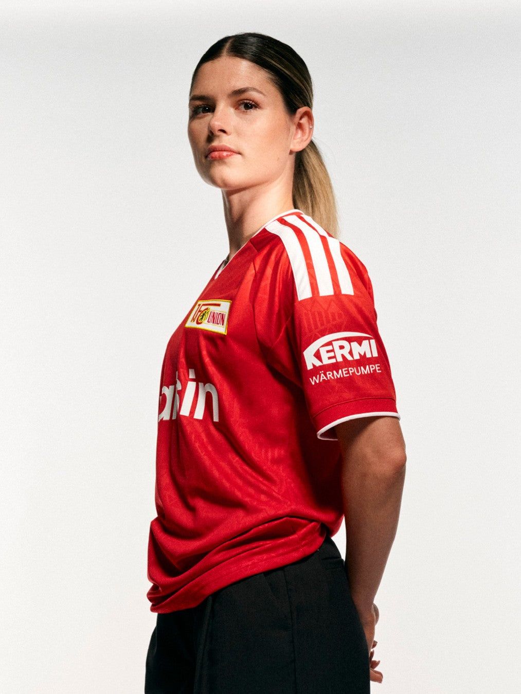 Eine Person trägt ein rotes Fußballtrikot mit weißen Streifen und steht in seitlicher Ansicht vor einem neutralen Hintergrund.
