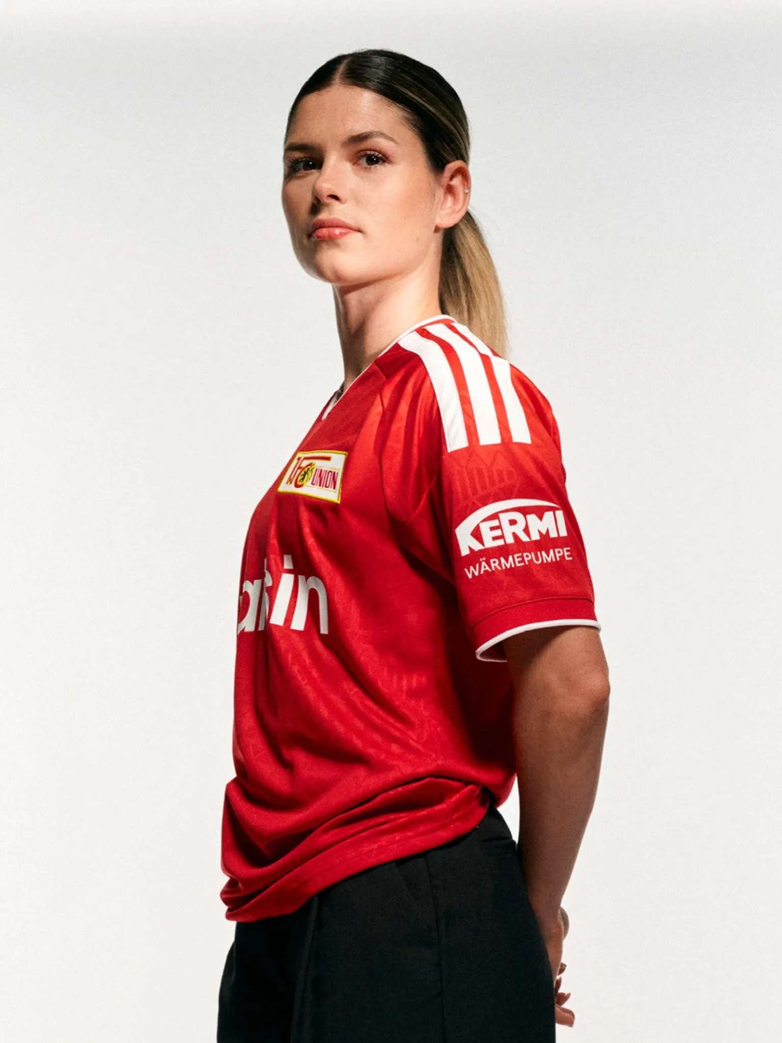 Eine Person trägt ein rotes Fußballtrikot mit weißen Streifen und steht in seitlicher Ansicht vor einem neutralen Hintergrund.