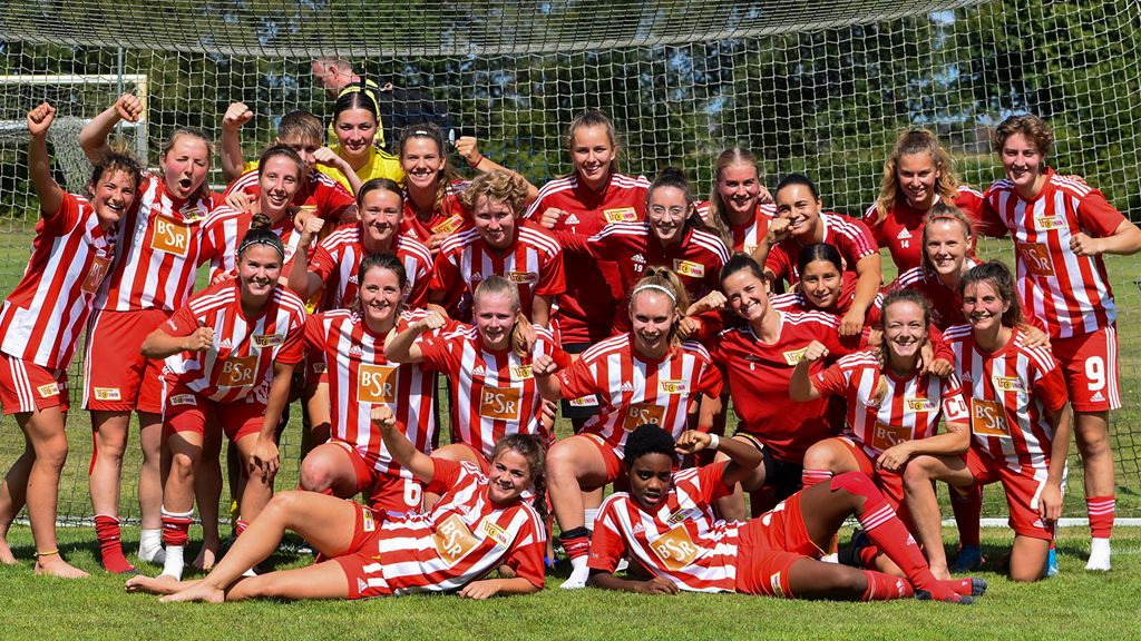 Eine Gruppe von Fußballspielerinnen in roten Trikots mit weißen Streifen. Sie freuen sich und lächeln.