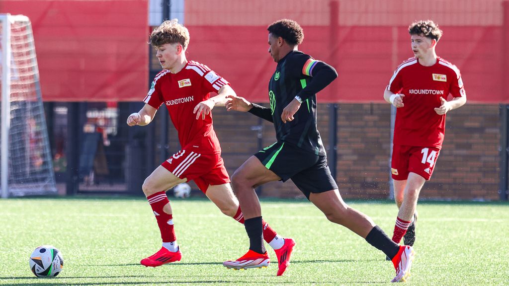 Drei Fußballspieler in action; zwei in roten Trikots und einer in schwarzem Trikot, auf einem Rasenplatz.