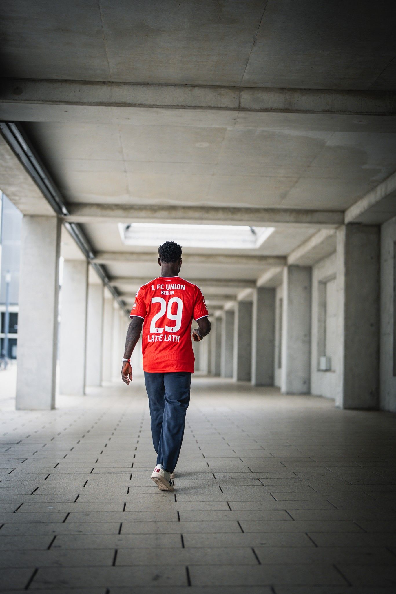 Person in rotem Fußballtrikot mit der Nummer 29 geht durch einen modernen, lichtdurchfluteten Gang.