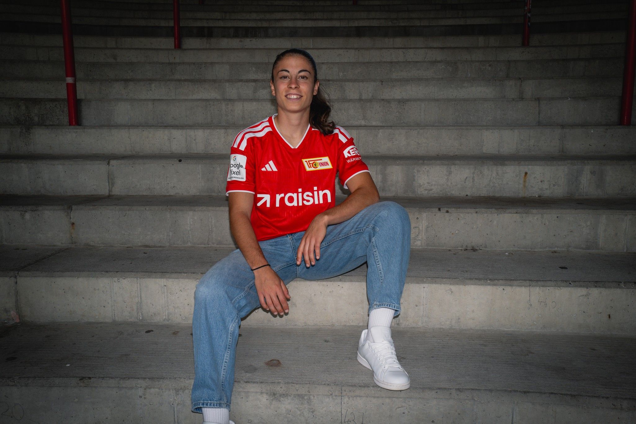 Eine Person in einem roten Fußballtrikot sitzt auf einer Treppe und trägt Jeans und weiße Sneakers.