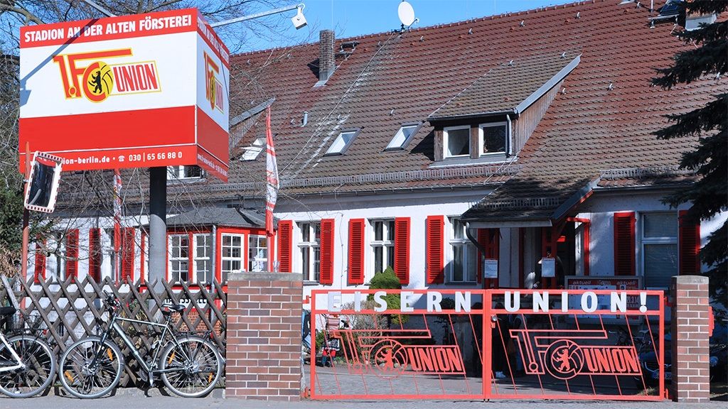 Ein Gebäude mit roter Fensterbekleidung, einem großen Schild mit "FC Union" und Fahrrädern vor einem Zaun.