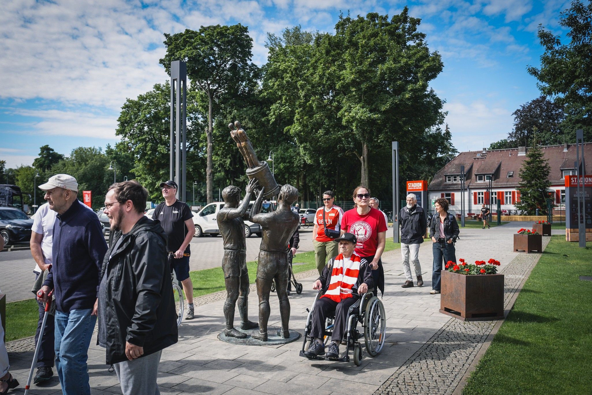 Menschen versammeln sich um eine Statue von zwei jubelnden Männern, während eine Person im Rollstuhl lächelt.