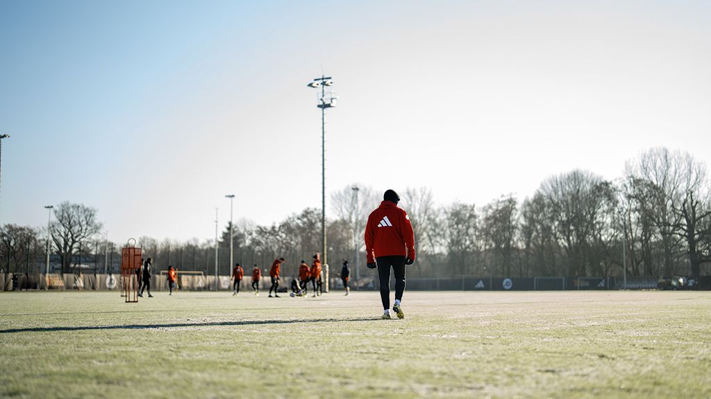 Ein Trainer in roter Jacke geht über ein Fußballfeld, während mehrere Spieler im Hintergrund trainieren.