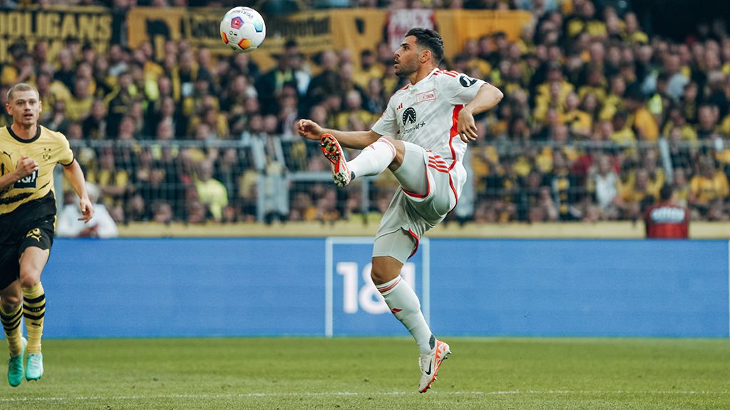 Ein Fußballspieler trägt ein graues Trikot. Er kickt den Ball hoch in die Luft. Die Zuschauer freuen sich.