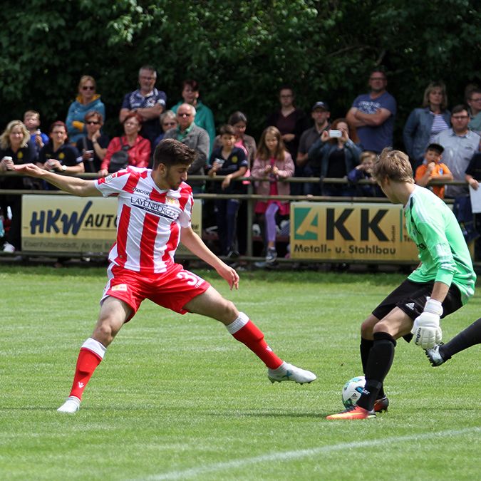 Ein Fußballspieler in rot-weißem Kleid versucht, den Ball zu schießen. Der Torwart von der anderen Mannschaft steht bereit. Viele Leute schauen zu.