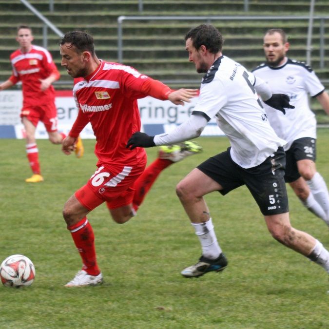 Zwei Fußballspieler in rot und schwarz jagen den Ball auf einem Rasenplatz, während sie sich um die Kontrolle bemühen.