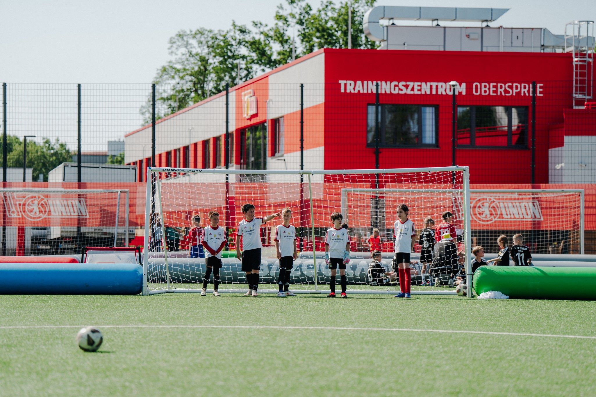 Sechs Kinder in Fußballtrikots stehen vor einem Tor auf einem Sportplatz, im Hintergrund ein rotes Trainingszentrum.