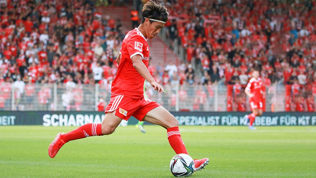 A soccer player in a red jersey kicks the ball during a game on a stadium field with cheering fans in the background.