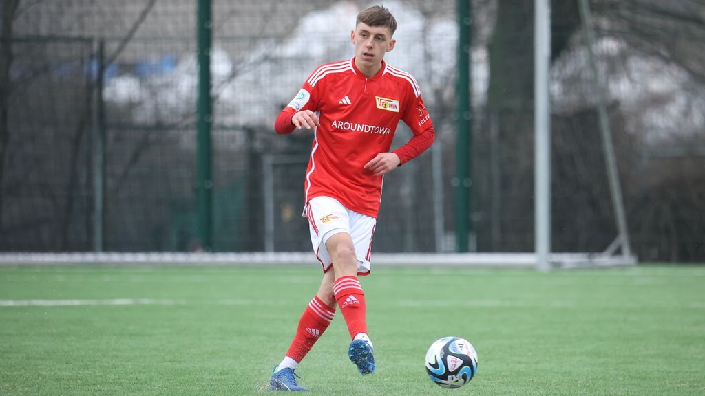 Ein junger Fußballspieler in rotem Trikot mit Vereinslogo dribbelt einen Ball auf einem Kunstrasenplatz.