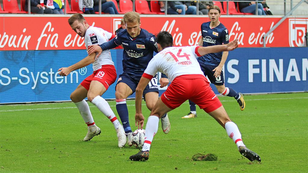 Zwei Fußballspieler kämpfen um den Ball, während ein dritter Spieler in der Nähe steht. Stadionatmosphäre im Hintergrund.