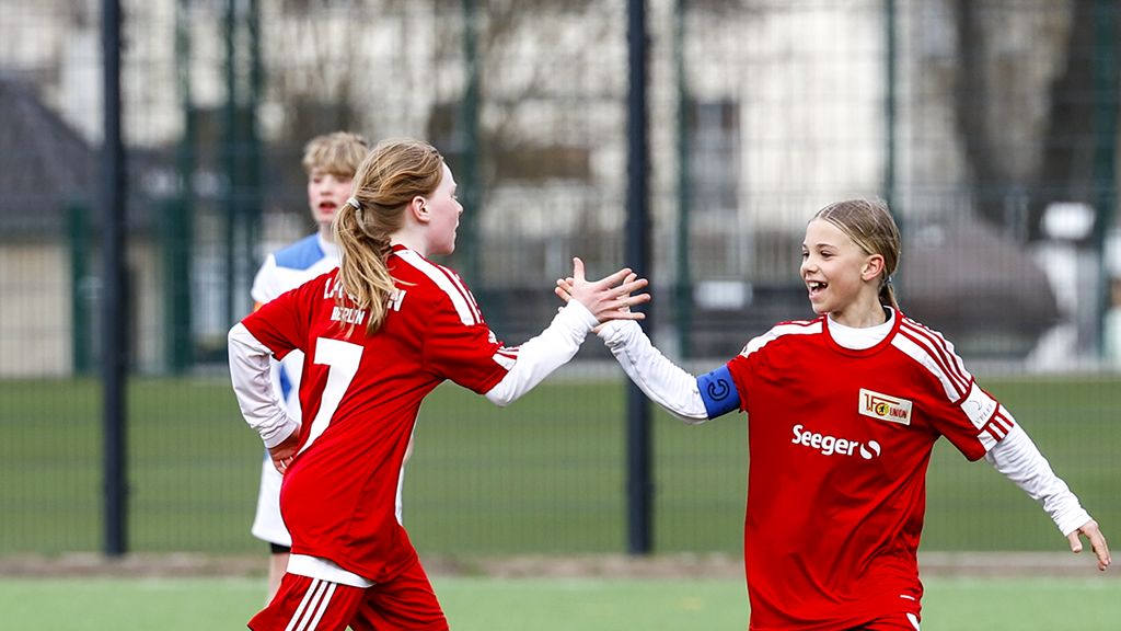 Zwei junge Fußballspielerinnen in roten Trikots feiern gemeinsam auf dem Spielfeld. Im Hintergrund steht ein weiteres Mädchen.