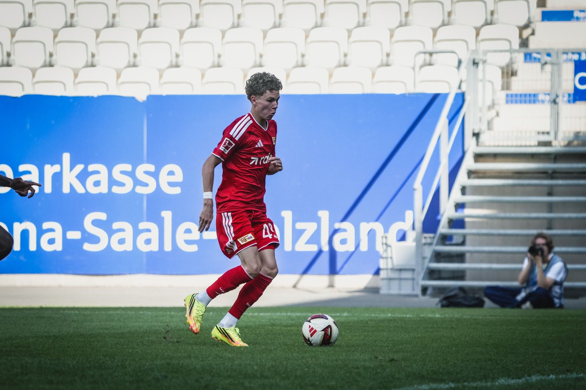 Ein Fußballspieler in rotem Trikot dribbelt den Ball auf einem Rasenfeld, im Hintergrund ist eine Tribüne zu sehen.
