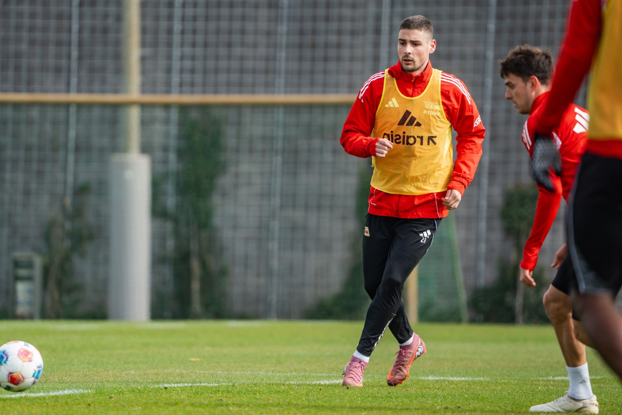 Zwei Fußballspieler trainieren auf einem Rasenplatz, einer trägt eine gelbe Trainingsweste, der andere ein rotes Shirt.