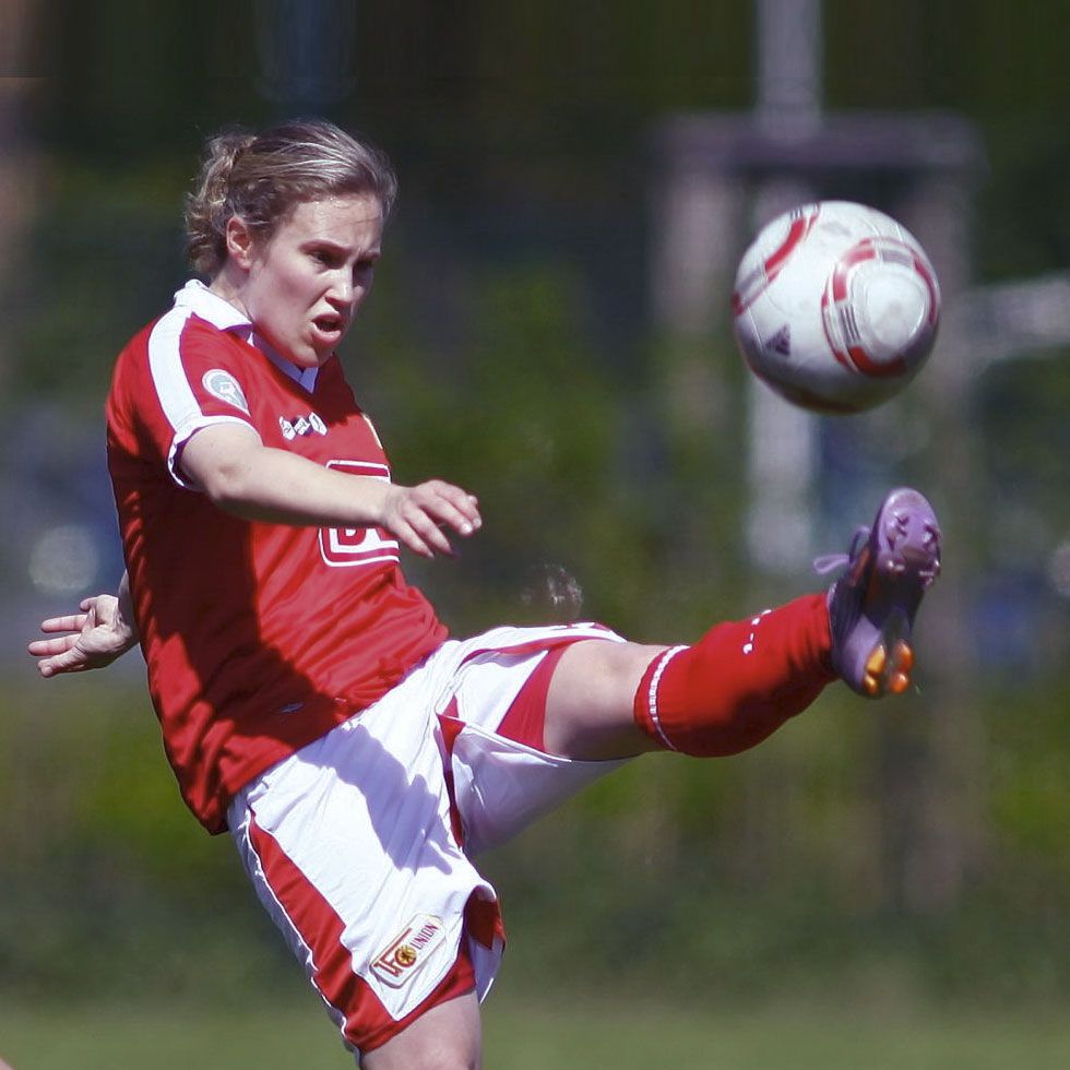 Eine Fußballspielerin in rotem Trikot hebt das Bein, um den Ball zu kicken, während sie konzentriert auf das Spiel schaut.