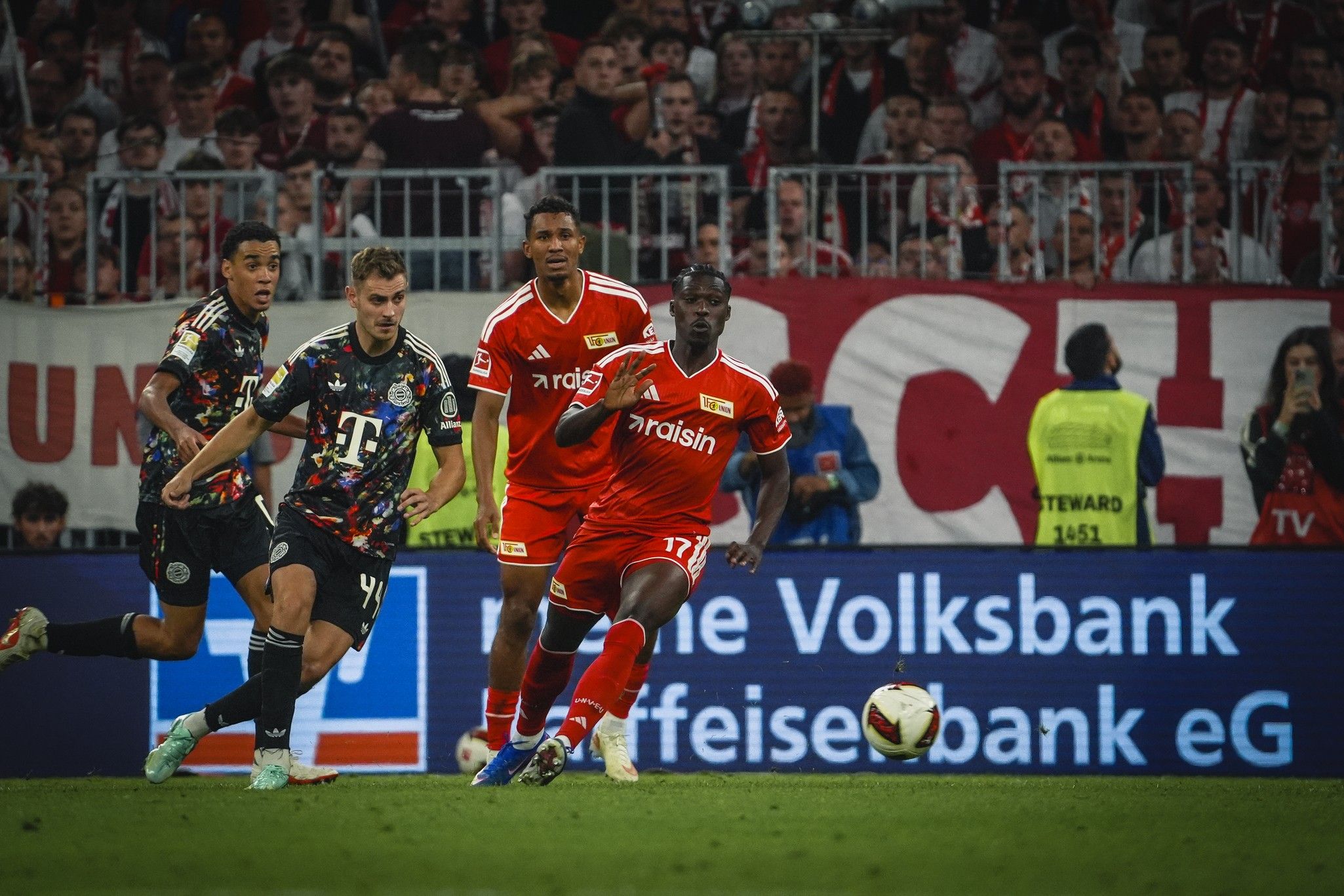 Spielszene im Fußball, Spieler von Union Berlin in roten Trikots, gegen Bayern München in bunten Trikots, Publikum im Hintergrund.