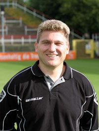 Ein lächelnder Mann in einem schwarzen Poloshirt steht im Freien, hinter ihm sind Sportinfrastruktur und Bäume sichtbar.