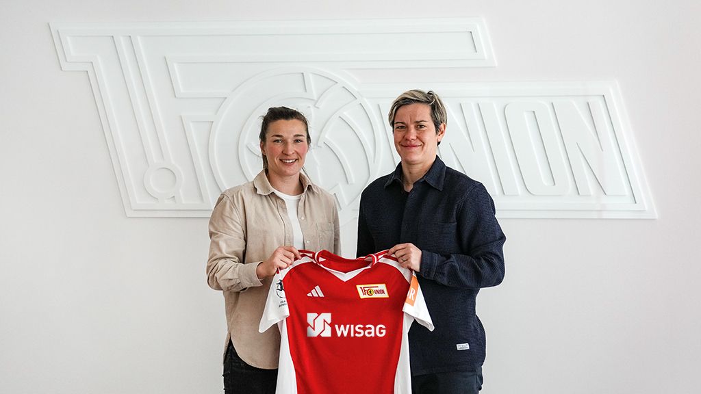 Zwei Menschen halten ein Trikot von der Fußballmannschaft 1. FC Union Berlin. Das Trikot ist vor einem Logo.